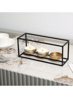 two candles are sitting in a metal holder on a marble countertop next to a cup and saucer