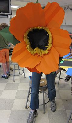 a woman standing in front of a large orange flower on top of a table with other people sitting at desks behind her