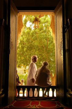 two people sitting on a bench looking out an open door at the trees and buildings