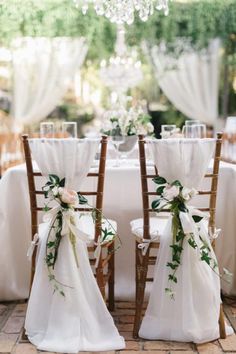 two wooden chairs decorated with white flowers and greenery sit in front of a table