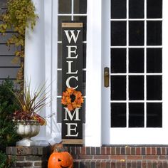 a welcome sign on the front door of a house with two pumpkins and flowers