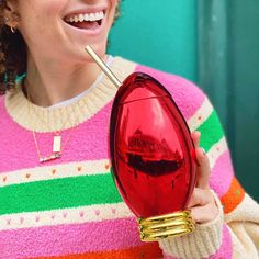 a smiling woman holding a red object in her hand with a straw sticking out of it's mouth