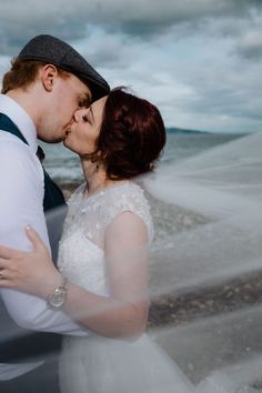 a bride and groom kissing on the beach with veil blowing in the wind behind them