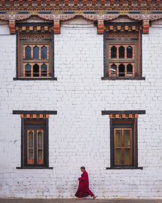 a woman in a long red dress walking past a white brick building with two windows