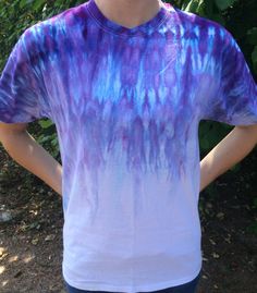 a man wearing a tie dyed shirt standing in front of some bushes and trees with his hands on his hips