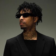 a man wearing sunglasses and a black suit with tattoos on his chest, standing in front of a gray background