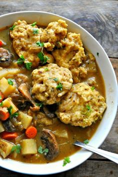 a white bowl filled with meat and vegetable stew on top of a wooden table next to a spoon