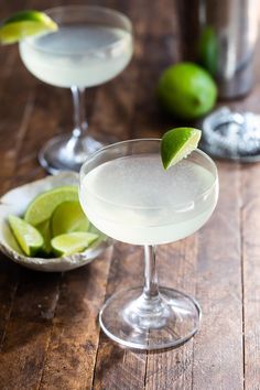 two margaritas with lime wedges in them on a wooden table next to a cocktail shaker