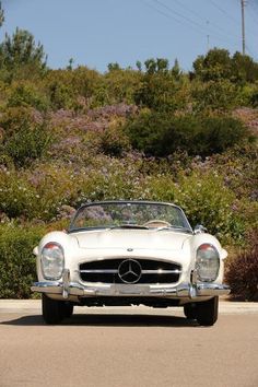 an old white mercedes sports car driving down the road in front of bushes and trees