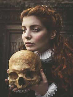 a woman with red hair holding a skull in front of her face and looking at the camera