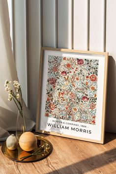 there is a vase with flowers on the table next to a framed print that says william morris