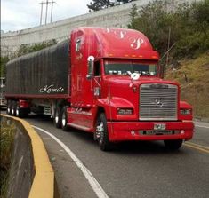 a red semi truck driving down a road next to a wall and trees on the side of the road