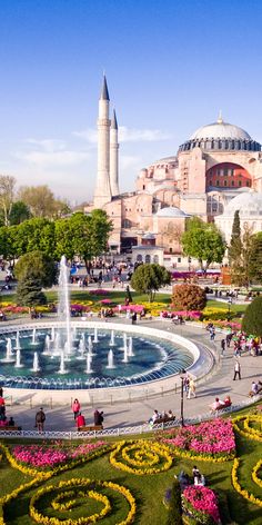 people are walking around in front of a fountain with flowers on the ground and buildings in the background