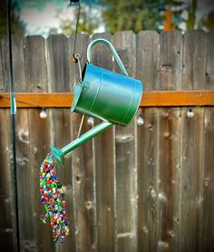 a green watering can filled with beads hanging from it's side next to a wooden fence