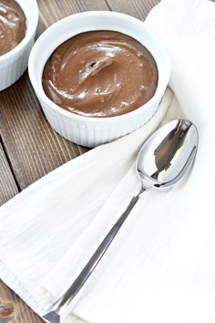 two white bowls filled with chocolate pudding on top of a wooden table next to a spoon