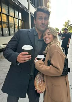 a man and woman standing next to each other on the street holding coffee cups in their hands