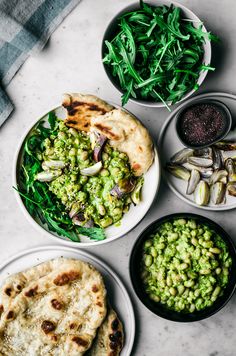 four plates filled with different types of food on top of a white tablecloth next to bowls of vegetables and pita bread