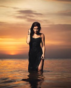 a woman standing in the water at sunset with her hand on her head and wearing a black dress