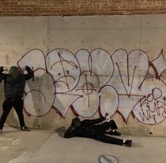 two people standing next to a wall with graffiti on it and one person laying down