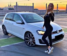 a woman standing next to a white car in an empty parking lot with the sun setting behind her