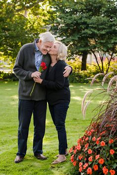 an older couple embracing each other while standing in the grass with red flowers behind them