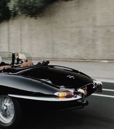 an old black sports car driving down the road with two people in it's seat