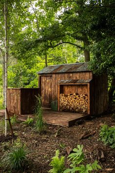 an outhouse in the woods with firewood stacked on it's sides and trees around