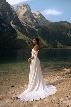 a woman in a long white dress standing on the shore of a lake with mountains in the background