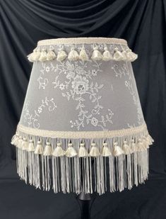 a white lamp shade with tassels and beads on the bottom, sitting on a black table