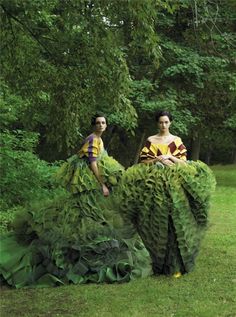 two women in dresses made out of leaves sitting on top of each other with trees behind them