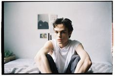 a young man sitting on top of a bed next to a white wall in a bedroom