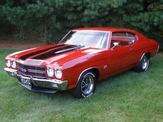 an old red car is parked in the grass near some trees and bushes with black stripes on it's hood