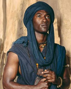 a man wearing a blue turban standing in front of a stone wall with his hands crossed