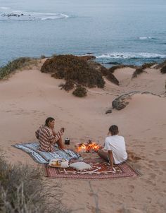 two people sitting on a blanket at the beach eating food and drinking beer by the fire