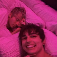 two young men laying in bed with pink lights on the head and one is smiling