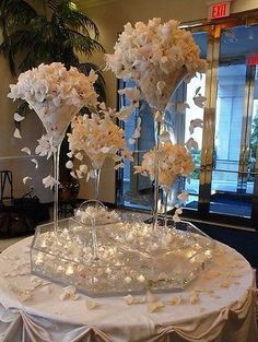 three tall clear vases with flowers on top of a white table cloth covered round table