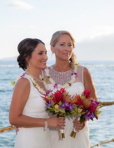 two women standing next to each other holding bouquets in front of the ocean on a sunny day