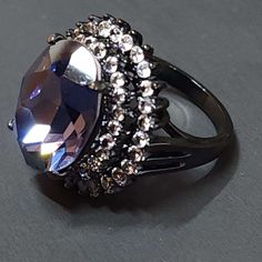 "Add an edgy vibe to your look with our Purple and Black Goth Ring. This statement piece features one large purple (Tanzanite) faceted CZ surrounded with many small clear faceted CZs cubic zirconia set against a black backdrop for a striking contrast. The cocktail ring is designed to make a statement with its unique and intricate details that exude boldness and confidence. Whether you're dressing up for a night out or want to elevate your casual look, our Purple and Black Goth Ring will be the perfect finishing touch to any outfit. Get ready to turn heads and receive compliments with this must-have addition to your jewelry collection. This is a high quality ring, Goth jewelry. Statement ring. Lots of sparkle. The front of the ring is 24mm tall.  The ring band is 2.5mm wide. The purple ston Elegant Black Amethyst Promise Ring, Gothic Party Ring Jewelry, Gothic Party Ring, Elegant Black Crystal Ring For Party, Elegant Purple Crystal Ring For Party, Elegant Purple Crystal Party Ring, Christian Jewelry Necklaces, Black Statement Ring, Purple Tanzanite