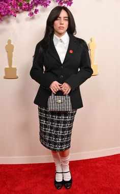 a woman in a skirt and jacket standing on a red carpet with purple flowers behind her