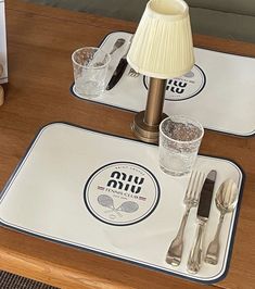 two place mats with silverware on top of a wooden table next to a lamp