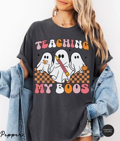 Get ready for a spooky season in style with this Comfort Colors® Teacher Halloween Shirt. This Retro Ghost Teacher Shirt, featuring the fun "Teaching My Boos" design, is the perfect Halloween Teacher Shirt for any classroom, making it a must-have Spooky Teacher T-shirt. Comfort Colors® Teacher Halloween Shirt, Halloween Teacher Shirt, Teaching My Boos Shirt, Spooky Teacher T-shirt, Retro Ghost Teacher Shirt About Comfort Colors C1717 The Models in the pictures are wearing 1 or 2 sizes up, Please Casual Halloween Shirt With Graphic Design, Short Sleeve Halloween Graphic Shirt, Casual Halloween Graphic Tops, Casual Graphic Tops For Halloween, Halloween Graphic Tee Shirt With Graphic Design, Halloween Graphic Tee With Graphic Design, Funny Graphic Print Shirt For Fall, Fall Graphic Tee Shirt With Character Print, Fall Graphic Tee With Character Print