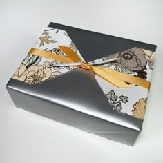 a silver box with a yellow ribbon tied around the edge and flowers on it, sitting on a white surface