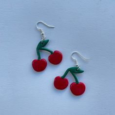 two cherries are hanging from silver earwires on a white surface, one is red and the other is green