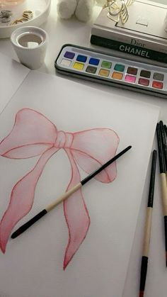 watercolors and brushes are sitting on a table next to a drawing of a pink bow