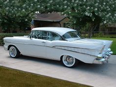 an old white car is parked in front of a house with flowers on the driveway