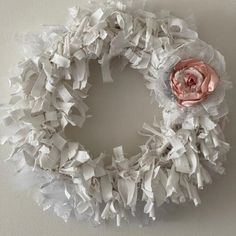 a white wreath with pink flower on top of it and ribbon around the edges, hanging from a wall