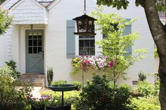 a bird bath in front of a white house with blue shutters and pink flowers