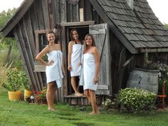 three women in white robes standing outside of a wooden building with an outhouse behind them