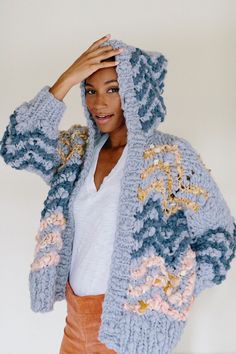 a woman wearing a blue crochet jacket and orange pants with her hands on her head