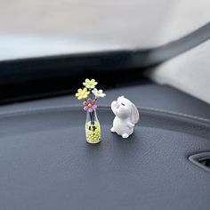 a small white rabbit next to a yellow vase with flowers in it on top of a car dashboard